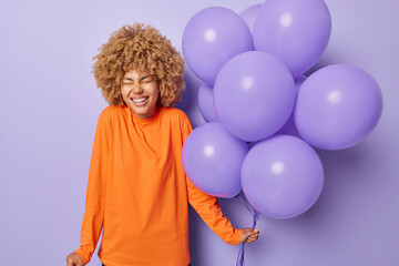 Wall Mural - Positive curly haired young woman has festive mood dressed in casul orange jumper holds bunch of inflated balloons laughs happily isolated over purple background. People and holiday concept.