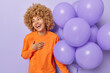 Attractive curly haired woman laughs joyfully keeps hand on chest looks away glad to receive congratulations and warm words holds big bunch of balloons wears orange jumper isolated on purple wall