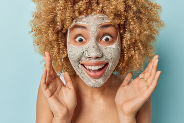 Wall Mural - Portrait of surprised cheerful woman with curly blonde hair keeps palms raised stares shocked at camera smiles broadly undergoes beauty procedures applies clay mask isolated over blue background