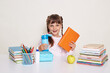 © sementsova321 - Horizontal shot of winsome positive little schoolgirl with dark hair and braids, holding book and blue water bottle, sitting at table surrounded with books against white wall.