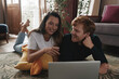 © gstockstudio - Beautiful young couple using laptop and smiling while lying on the floor at home together