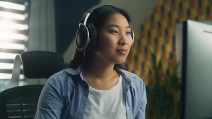 Poster - Asian woman in headphones sitting at the table at home and browsing the internet on computer on weekend or working on project remotely