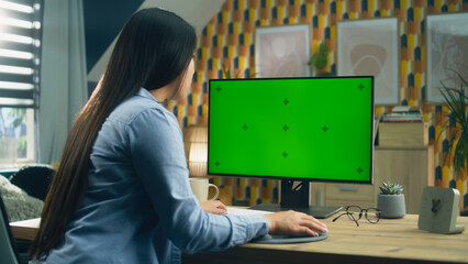 Poster - Asian woman sitting at the table and looking at screen of personal computer while surfing the internet in spare time or working remotely from home. Freelance. Green screen and chromakey