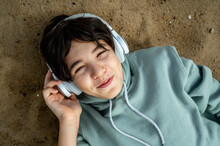 Smiling Boy Wearing Wireless Headphones Listening To Music And Relaxing On Sand