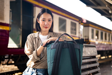 Happy beautiful asian backpacker woman wearing jacket and backpack enjoys moment of success with his holiday, she smiling and sitting in the train station going to travel.