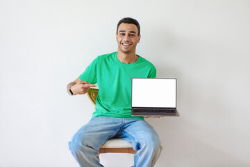 Poster - Happy arab man pointing at laptop pc with empty screen, sitting on chair against light studio wall, mockup for website
