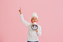 News. Studio Shot Of Little Girl, Kid In Casual Style Clothes And Warm Hat Shouting At Megaphone Isolated Over Pink Background.