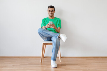 Wall Mural - Excited young arab man using cellphone and texting or networking in social media sitting on chair over light wall