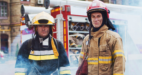 Medium plan of two firefighters in helmet and gull equipment standing next to car and looking at camera. Smoke from the fire covers rescuers and a fire truck. Concept of saving lives, fire safety