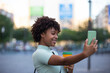 © jcalvera - making selfie. Young woman with curly hair wearing fashionable clothes outdoors in the city.