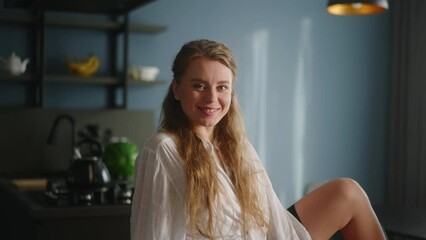 Wall Mural - Attractive woman looking at camera sitting on the couch on the dark modern stylish kitchen background. Close-up face portrait of young smiling woman. Blonde long-haired female sitting on back of sofa