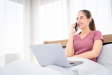 Fototapeta Psy - Asian woman sitting in a pink casual at home working sitting next to the bed Talking on the phone with a customer on a mobile phone work at home work happily relax