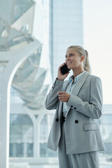 Poster - Happy young blond businesswoman in grey suit talking on mobile phone while standing by large iwndow in modern office