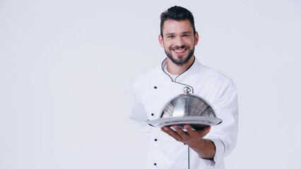 Wall Mural - smiling chef standing with hand behind back while holding serving dish with cloche isolated on white.