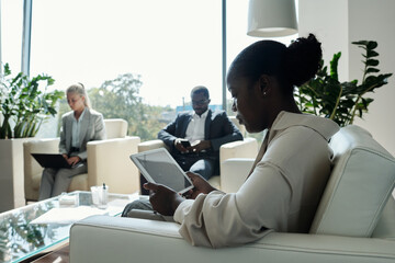 Aufkleber - Side view of young serious female office worker with tablet sitting on white leather couch in openspace office against two coworkers