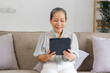 © NanSan - Happy mature asian woman using digital tablet looking at computer virtual calling having online meeting, sitting on couch.
