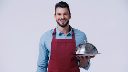 Wall Mural - smiling waiter in apron holding serving dish with cloche isolated on grey.