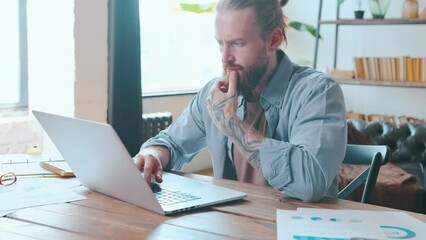Young focused brutal Caucasian man millennial with tattoo on arm works as programmer typing code on laptop for new software or developing website for IT company sits at table in modern office.