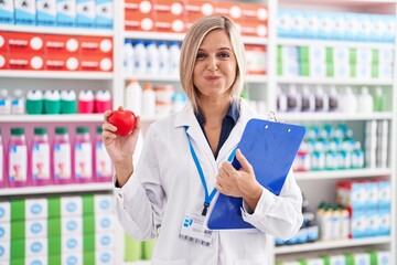 Wall Mural - Young woman working at pharmacy drugstore holding heart puffing cheeks with funny face. mouth inflated with air, catching air.