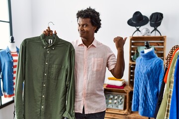 Poster - African man with curly hair holding shirt from clothing rack at retail shop pointing thumb up to the side smiling happy with open mouth