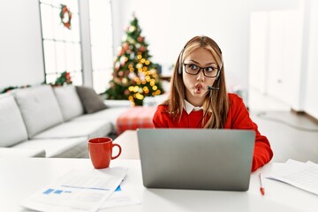 Poster - Young caucasian girl sitting on the table working using laptop by christmas tree making fish face with lips, crazy and comical gesture. funny expression.