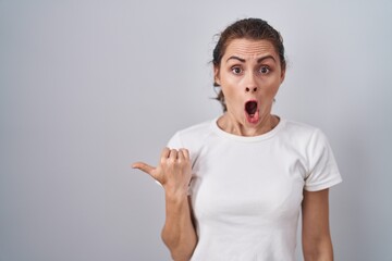 Poster - Beautiful brunette woman standing over isolated background surprised pointing with hand finger to the side, open mouth amazed expression.