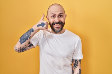 Wall Mural - Young hispanic man with tattoos standing over yellow background smiling doing phone gesture with hand and fingers like talking on the telephone. communicating concepts.