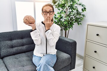 Canvas Print - Young hispanic woman holding brain working at psychology clinic covering mouth with hand, shocked and afraid for mistake. surprised expression