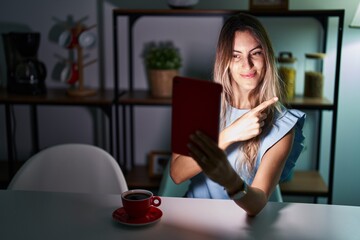 Canvas Print - Young hispanic woman using touchpad sitting on the table at night pointing with hand finger to the side showing advertisement, serious and calm face