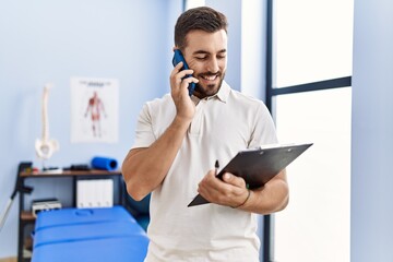 Poster - Young hispanic man wearing physiotherapist uniform talking on the smartphone at clinic