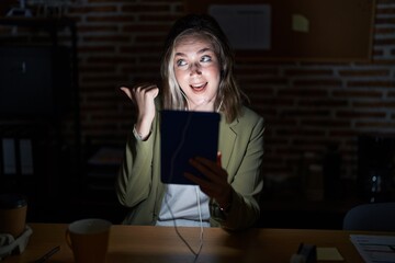 Poster - Blonde caucasian woman working at the office at night smiling with happy face looking and pointing to the side with thumb up.