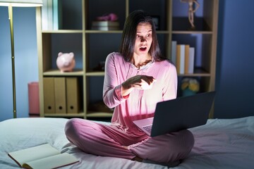 Sticker - Young hispanic woman using computer laptop on the bed pointing with finger surprised ahead, open mouth amazed expression, something on the front