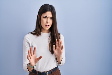 Poster - Young brunette woman standing over blue background moving away hands palms showing refusal and denial with afraid and disgusting expression. stop and forbidden.