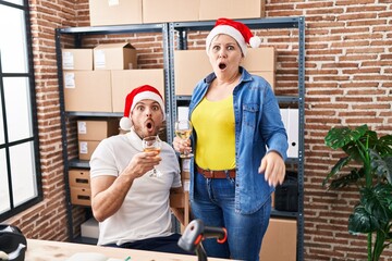 Wall Mural - Hispanic man and woman working at small business ecommerce drinking wine at christmas scared and amazed with open mouth for surprise, disbelief face