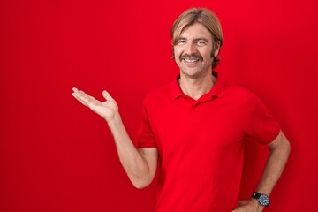 Sticker - Caucasian man with mustache standing over red background smiling cheerful presenting and pointing with palm of hand looking at the camera.