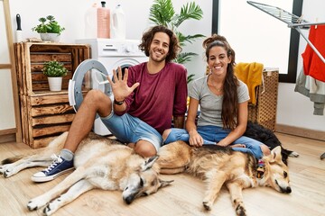 Wall Mural - Young hispanic couple doing laundry with dogs showing and pointing up with fingers number five while smiling confident and happy.