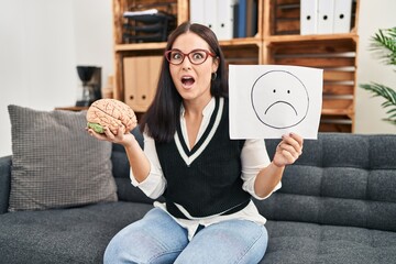 Wall Mural - Young hispanic woman working on depression holding sad emotion paper and brain afraid and shocked with surprise and amazed expression, fear and excited face.
