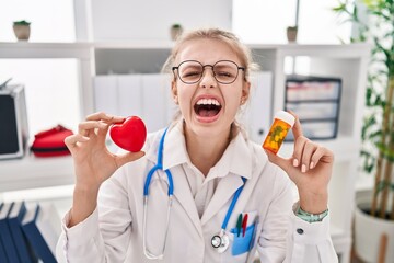 Wall Mural - Young caucasian doctor woman holding pills and heart angry and mad screaming frustrated and furious, shouting with anger looking up.