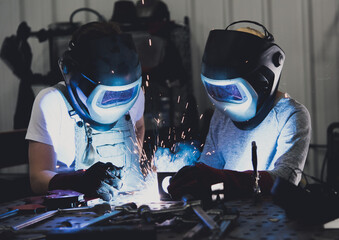 Industrial woman welder teaches younger student how to MIG weld metal with a torch