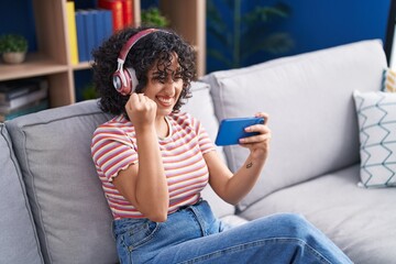 Sticker - Young middle eastern woman playing video game sitting on sofa at home