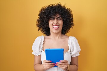 Poster - Young brunette woman with curly hair using touchpad over yellow background sticking tongue out happy with funny expression.