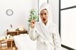 © Krakenimages.com - Young brunette woman wearing towel and bathrobe standing at beauty center pointing finger up with successful idea. exited and happy. number one.
