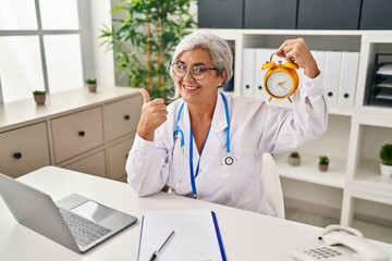 Poster - Middle age woman with grey hair wearing doctor uniform holding alarm clock smiling happy and positive, thumb up doing excellent and approval sign