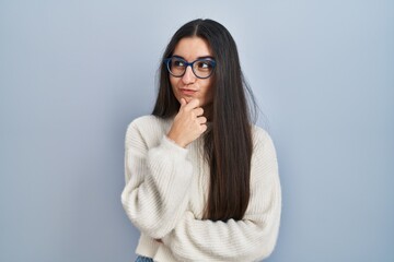 Poster - Young hispanic woman wearing casual sweater over blue background looking confident at the camera with smile with crossed arms and hand raised on chin. thinking positive.