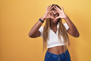 Poster - African american woman with braided hair standing over yellow background doing heart shape with hand and fingers smiling looking through sign