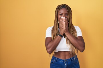 Poster - African american woman with braided hair standing over yellow background laughing and embarrassed giggle covering mouth with hands, gossip and scandal concept