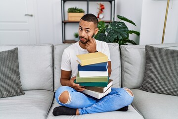 Poster - African american young man holding a pile of books sitting on the sofa pointing to the eye watching you gesture, suspicious expression