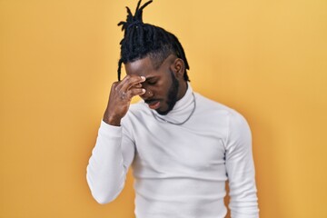 Wall Mural - African man with dreadlocks wearing turtleneck sweater over yellow background tired rubbing nose and eyes feeling fatigue and headache. stress and frustration concept.