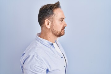 Poster - Middle age caucasian man standing over blue background looking to side, relax profile pose with natural face and confident smile.