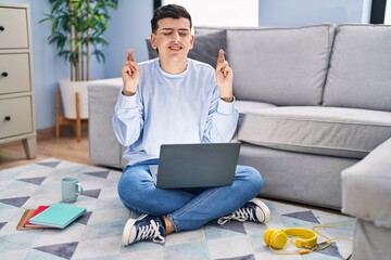 Wall Mural - Non binary person studying using computer laptop sitting on the floor gesturing finger crossed smiling with hope and eyes closed. luck and superstitious concept.